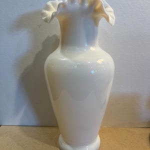 May include: A white milk glass vase with a smooth, bulbous body and a flared, ruffled rim. The vase features a glossy finish and a pedestal base, standing upright with a narrow neck that widens toward the top opening.