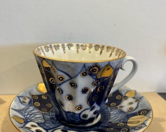 Może przedstawiać: Zestaw filiżanki ze spodkiem z porcelany w głębokim niebiesko-białym wzorze ze złotymi akcentami. Projekt zawiera małe motywy kwiatowe, geometryczne kształty i złocony brzeg. Filiżanka ma zakrzywione ucho i żłobkowany kształt korpusu.