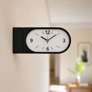 May include: A wall-mounted clock with a matte black rectangular base and an oval-shaped face. The white clock face features black numerals at 3, 6, 9, and 12 o'clock, with simple black tick marks for the remaining hours and black hands.