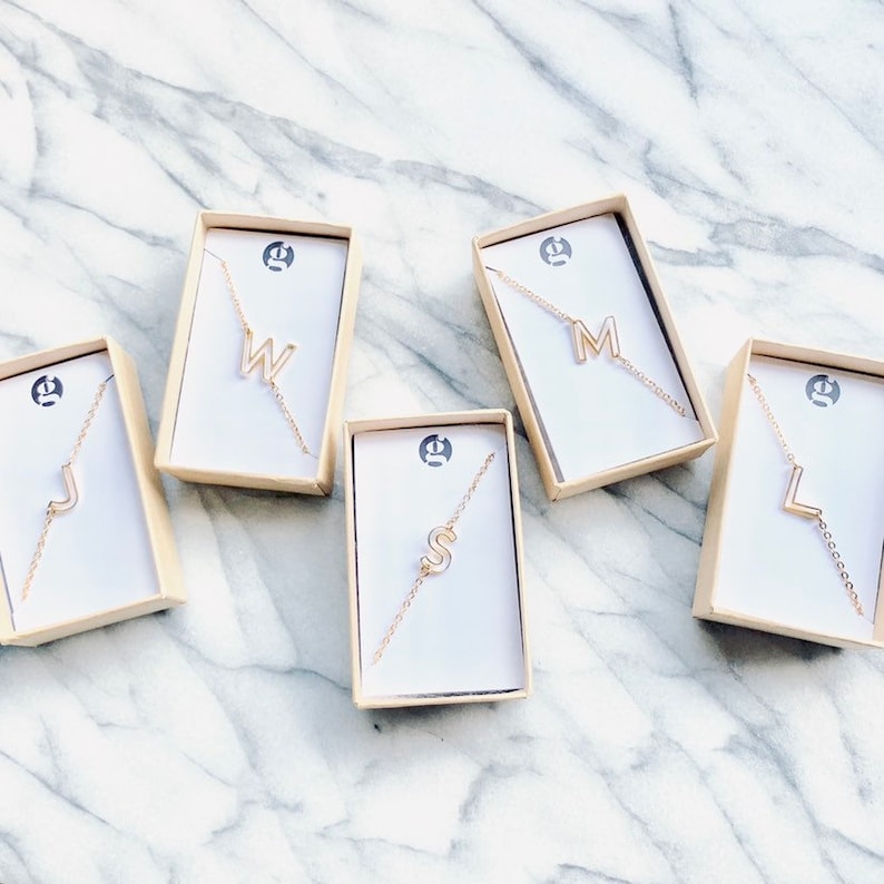 May include: Five small white boxes with gold trim, each containing a gold chain necklace with a different letter pendant. The letters are J, W, M, S, and L.
