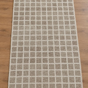 Puede incluir: Una alfombra rectangular que presenta un patrón de cuadrícula repetitivo de líneas beige claro sobre un fondo taupe texturizado. El diseño consiste en cuadrados uniformes dispuestos en una estructura organizada por toda la superficie de la alfombra.