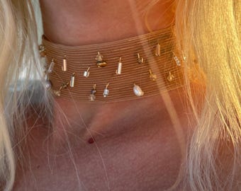 May include: A tan mesh choker necklace featuring scattered metallic cylindrical beads and small white pearls. The wide, flexible band sits snugly around the neck, with the beads and pearls attached to the fine, horizontal wire-like mesh structure.