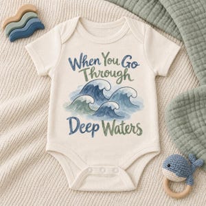 May include: A cream-colored short-sleeve baby bodysuit featuring a watercolor illustration of blue and green ocean waves. The text "When You Go Through Deep Waters" is printed in a dark blue and green script font across the chest area.