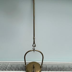 Op de afbeelding: Een vintage koperen hangende olielamp met een ronde voet, een centraal brandermechanisme, een gebogen handvat aan de bovenkant en een lange verticale stang die eindigt in een haak voor ophanging. Het metaal heeft een verouderde patina.