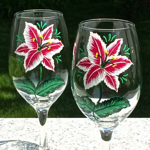 This is a set of two hand painted wine glasses. Each glass has a single red and white lily with green leaves. Light green wisps surround the flowers. Each glass has a red and white beaded wine glass charm attached to the base.