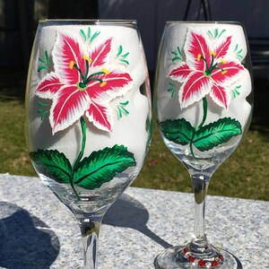 This is a set of two hand painted wine glasses. Each glass has a single red and white lily with green leaves. Light green wisps surround the flowers. Each glass has a red and white beaded wine glass charm attached to the base.