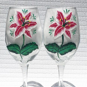 This is a set of two hand painted wine glasses. Each glass has a single red and white lily with green leaves. Light green wisps surround the flowers. Each glass has a red and white beaded wine glass charm attached to the base.