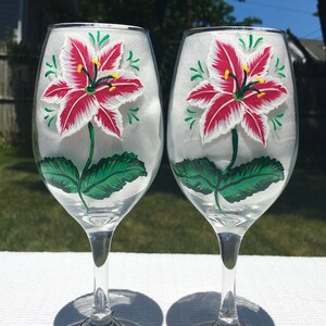 This is a set of two hand painted wine glasses. Each glass has a single red and white lily with green leaves. Light green wisps surround the flowers. Each glass has a red and white beaded wine glass charm attached to the base.