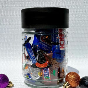 Pu&ograve; includere: Un barattolo di vetro trasparente con coperchio nero, riempito con barrette di cioccolato assortite tra cui Crunch e Snickers. L'esterno del barattolo presenta sagome di pipistrelli neri e un adesivo a forma di cappello da strega.