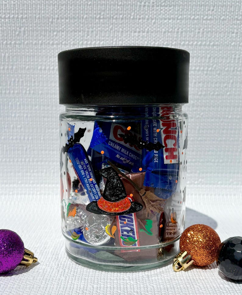 Pu&ograve; includere: Un barattolo di vetro trasparente con coperchio nero, riempito con barrette di cioccolato assortite tra cui Crunch e Snickers. L'esterno del barattolo presenta sagome di pipistrelli neri e un adesivo a forma di cappello da strega.