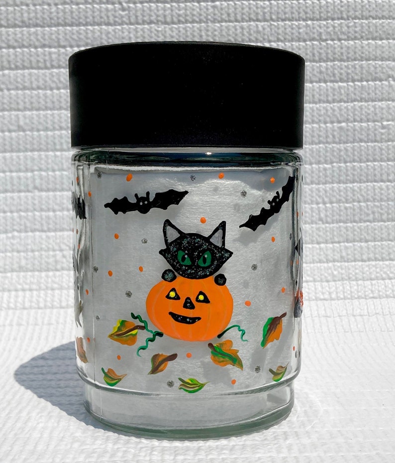 Pu&ograve; includere: Un barattolo di vetro trasparente con coperchio nero decorato con un motivo di Halloween dipinto a mano. Il disegno raffigura un gatto nero con occhi verdi seduto su una zucca arancione, circondato da pipistrelli neri, puntini e foglie autunnali.
