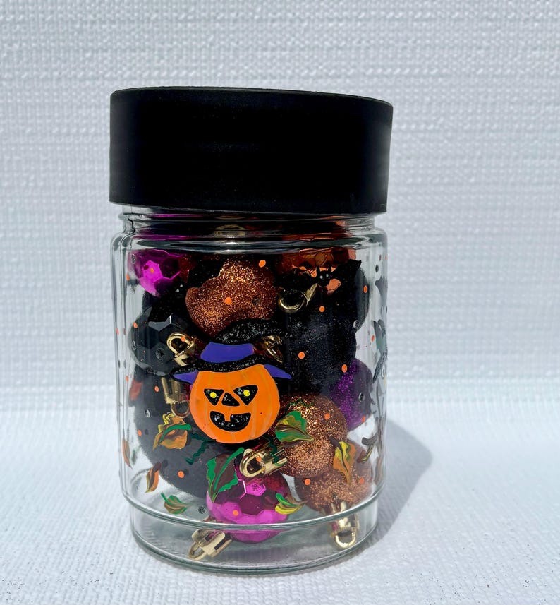 Pu&ograve; includere: Un barattolo di vetro trasparente con coperchio nero riempito con piccole sfere decorative assortite in nero, arancione e viola. Il barattolo presenta un volto di zucca dipinto in arancione con un cappello viola e dettagli di foglie verdi.