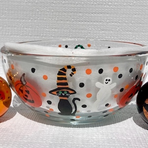 May include: A clear glass bowl decorated with painted Halloween motifs including a black cat wearing a striped witch hat, a white ghost, and orange pumpkins, surrounded by black and orange polka dots, flanked by two jack-o'-lantern ornaments.
