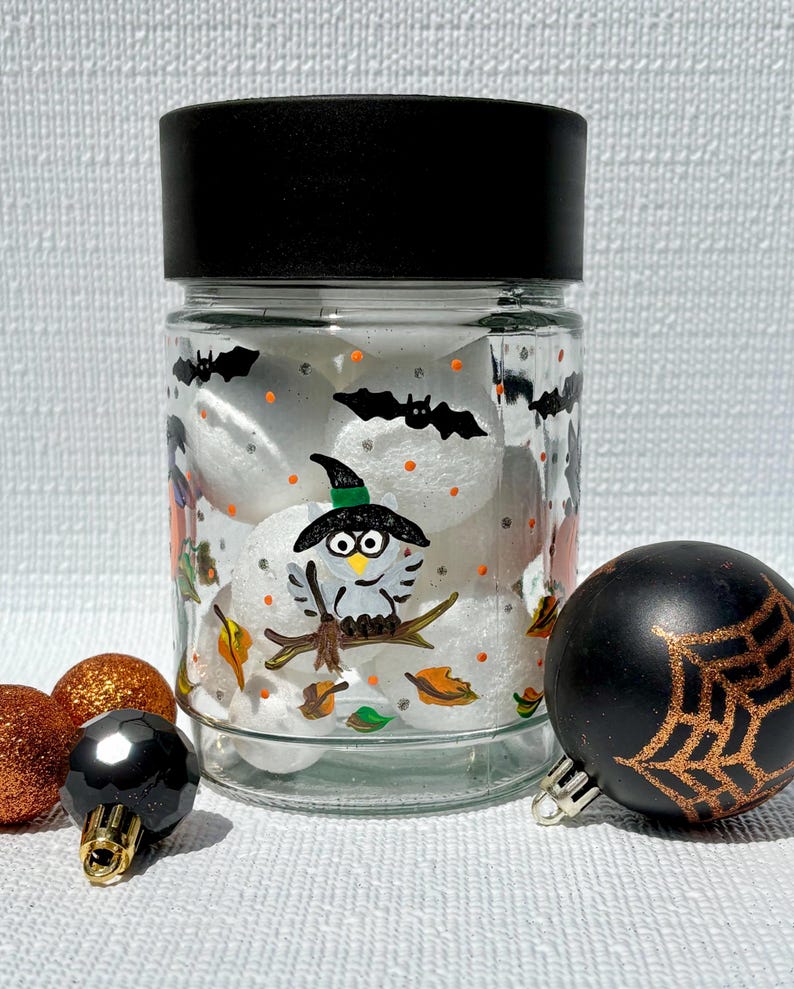 Pu&ograve; includere: Un barattolo di vetro trasparente con coperchio nero, riempito con oggetti sferici bianchi. Il barattolo presenta motivi di Halloween dipinti a mano, tra cui un gufo con cappello da strega, pipistrelli neri, punti arancioni e foglie autunnali, con accenti di glitter argento.
