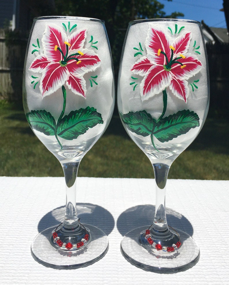 This is a set of two hand painted wine glasses. Each glass has a single red and white lily with green leaves. Light green wisps surround the flowers. Each glass has a red and white beaded wine glass charm attached to the base.