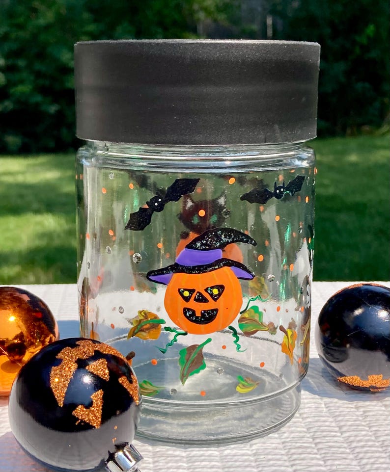 Pu&ograve; includere: Un barattolo di vetro trasparente con coperchio nero, decorato con motivi di Halloween dipinti a mano tra cui una zucca arancione centrale con un cappello a tesa viola, pipistrelli neri, foglie verdi e punti arancioni, con due decorazioni.
