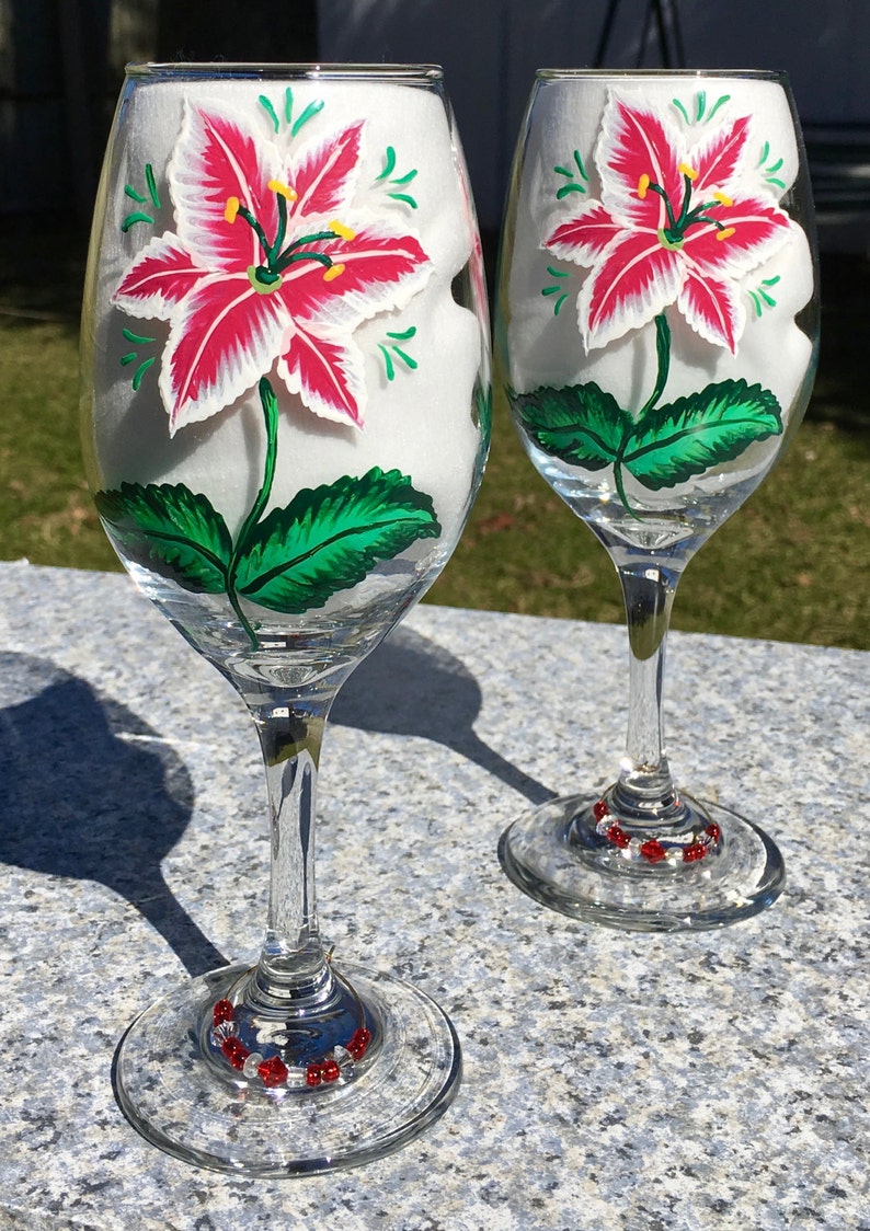 This is a set of two hand painted wine glasses. Each glass has a single red and white lily with green leaves. Light green wisps surround the flowers. Each glass has a red and white beaded wine glass charm attached to the base.