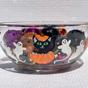 May include: A clear glass bowl decorated with hand-painted Halloween motifs including a black cat sitting inside an orange pumpkin, white ghosts, and small orange dots. The bowl is filled with various glittery spherical ornaments in purple, orange, and black.