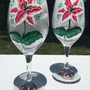 This is a set of two hand painted wine glasses. Each glass has a single red and white lily with green leaves. Light green wisps surround the flowers. Each glass has a red and white beaded wine glass charm attached to the base.