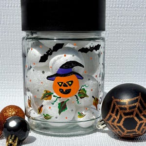Pu&ograve; includere: Un barattolo di vetro trasparente con coperchio nero, riempito con sfere di schiuma bianca. Il barattolo presenta disegni dipinti a mano di una zucca con un cappello viola, pipistrelli neri e foglie autunnali. Accanto al barattolo ci sono due piccoli ornamenti.