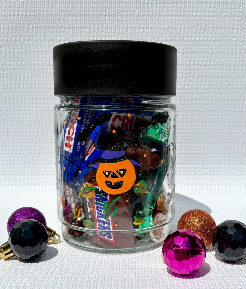 Pu&ograve; includere: Un barattolo di vetro trasparente con coperchio nero riempito di barrette di cioccolato assortite. La parte anteriore del barattolo presenta una zucca di Halloween arancione dipinta con un cappello da strega viola, circondata da foglie autunnali.