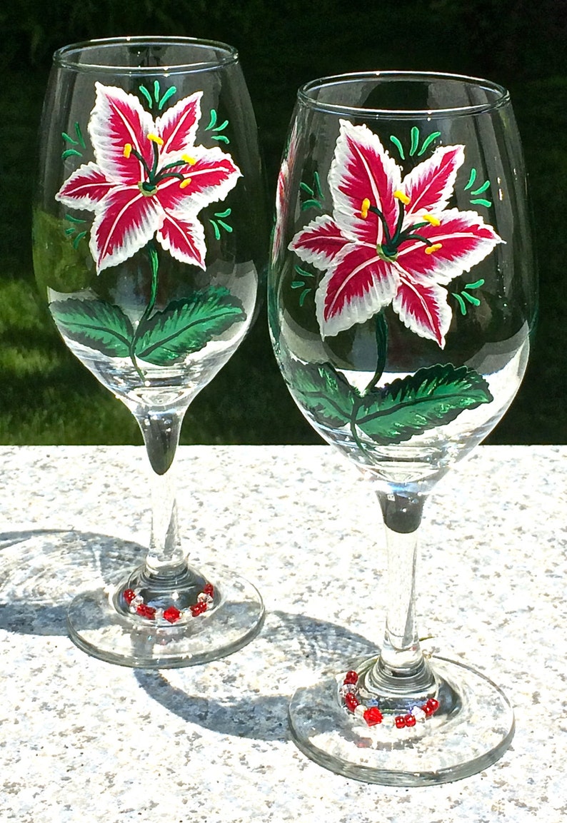 This is a set of two hand painted wine glasses. Each glass has a single red and white lily with green leaves. Light green wisps surround the flowers. Each glass has a red and white beaded wine glass charm attached to the base.