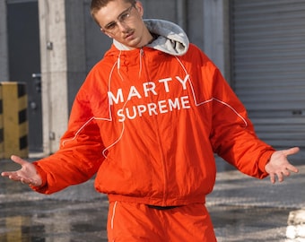 Peut inclure: Un coupe-vent zippé orange vif avec une doublure de capuche grise et le texte blanc MARTY SUPREME imprimé sur la poitrine. La veste présente de fins détails de passepoil blanc le long des manches et sur le devant, avec une fermeture éclair.