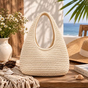 May include: A cream-colored, hand-crocheted hobo bag with a single integrated handle. The bag features a dense, horizontal stitch pattern throughout its rounded, triangular silhouette, providing a structured yet flexible form for carrying personal items.