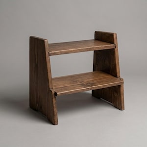 May include: A two-tier wooden step stool with a dark brown stained finish. The stool features two horizontal rectangular platforms supported by two vertical side panels that extend slightly above the top step, showcasing a visible wood grain.