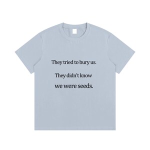 Könnte beinhalten: Hellblaues T-Shirt mit dem Text: „Sie versuchten, uns zu begraben. Sie wussten nicht, dass wir Samen waren.“ Der Text ist in einer einfachen Schriftart gehalten. Das T-Shirt hat einen Rundhalsausschnitt und kurze Ärmel.