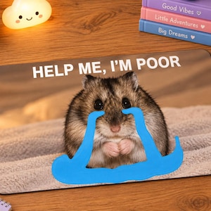 Op de afbeelding: Een rechthoekige muismat met een bruine en grijze hamster met grote zwarte ogen. De hamster heeft blauwe cartoon-tranen onder de ogen getekend. De tekst HELP ME, I'M POOR staat in witte hoofdletters bovenaan de muismat gedrukt.
