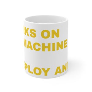 Può includere: Una tazza in ceramica bianca con testo giallo in grassetto che recita WORKS ON MACHINE, DEPLOY AND. La tazza ha una forma cilindrica con una finitura liscia e un manico standard, mostrando il testo stampato chiaramente sulla superficie bianca.