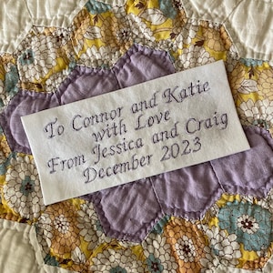 May include: A white fabric label with purple embroidery that reads "To Connor and Katie with Love from Jessica and Craig December 2023".