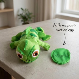 Op de afbeelding: Een felgroene pluche leguaan met een getextureerde rugkam en grote bruine ogen, vergezeld van een rond groen stoffen onderdeel met de tekst With magnetic suction cup en een pijl die naar het onderdeel wijst.