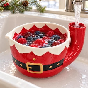 May include: A red and white ceramic bowl shaped like a Santa suit with a black belt and gold buckle. The bowl features a tall, cylindrical handle on the right side that acts as a water spout, currently being filled with water and fresh berries.