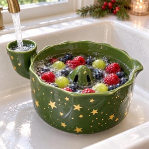 May include: A dark green ceramic berry colander with a gold star pattern, featuring a side spout for water intake and a central drainage cone. The bowl is filled with water, fresh red raspberries, and green grapes being rinsed under a tap.