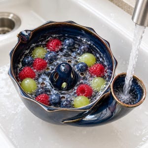 May include: A dark blue ceramic berry colander with a scalloped rim and a central drainage cone, filled with fresh red raspberries, blueberries, and green grapes being rinsed under a stream of water from a kitchen tap.