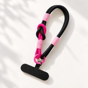 May include: A black and hot pink paracord lanyard featuring a knotted design, accented with light pink cord wrappings and a circular pink ring. Attached to the ring is a black, horizontal, T-shaped textured tab for securing items.