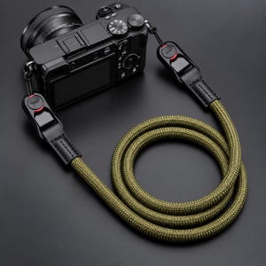 May include: A black digital camera attached to an olive green rope camera strap. The strap features black leather connectors with red accents at the attachment points. The rope is coiled in a circular pattern, showcasing a textured, woven design.