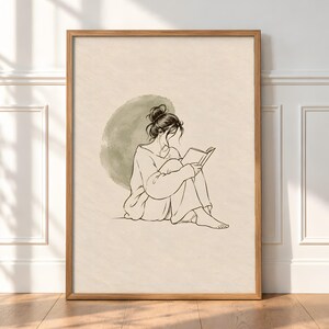 Puede incluir: Una lámina de arte enmarcada con un dibujo lineal de una persona con el pelo recogido en un moño, sentada y leyendo un libro. La figura está trazada en tinta negra sobre fondo crema con un suave círculo de acuarela verde oliva detrás.