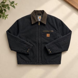 May include: A black canvas jacket featuring a brown corduroy collar, a full-length front zipper, and a zippered chest pocket. A square Carhartt brand patch is visible on the chest. The interior is lined with a green and brown plaid fabric.