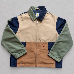 May include: A color-blocked Carhartt zip-up jacket featuring a tan upper chest, brown lower panels, navy blue side panels, and olive green sleeves. The jacket has a pointed collar, a full-length metal zipper, and a small Carhartt logo patch.