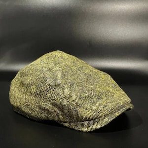 May include: A flat cap featuring a textured, woven fabric in a blend of olive green, yellow, and black threads. The cap has a rounded crown and a short, stiffened brim extending from the front, showcasing a dense, multi-coloured speckled pattern.