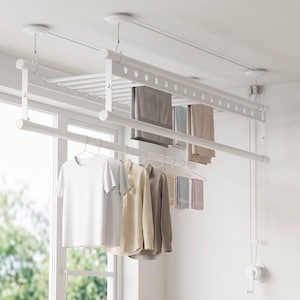 Könnte beinhalten: Ein weißer, an der Decke montierter Wäschetrockner mit Seilzugsystem und Handkurbel. Das Gestell verfügt über horizontale Stangen zum Aufhängen von Kleidung auf Bügeln und eine flache Ablagefläche, an der mehrere Kleidungsstücke hängen.