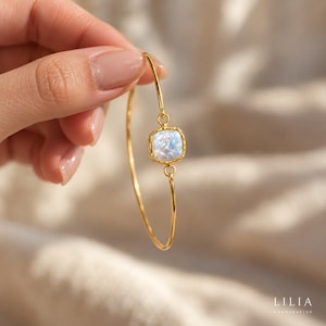 May include: A gold-toned bangle bracelet featuring a central square-shaped charm with a textured border and an iridescent, pearlescent inlay. The thin, polished metal band connects to the charm via small circular loops on either side.