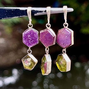 May include: Three silver pendants, each featuring two stacked hexagonal gemstones. The top stones are deep purple, while the bottom stones are a translucent olive green. Each pendant is held by a silver bail, showcasing a metallic setting.