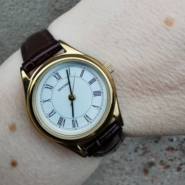 montre pour femme Sekonda vintage couleur or, cadran rond avec chiffres romains blancs et bracelet en cuir marron bordeaux.