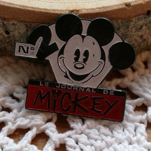Puede incluir: Un pin de esmalte metálico que muestra la cabeza de Mickey Mouse en blanco y negro. El pin incluye el texto N°2 y una base rectangular roja con el texto negro LE JOURNAL DE MICKEY, con un pequeño icono de la cabeza de Mickey sobre la letra I.