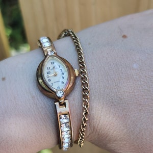 May include: A gold-coloured quartz watch with a white face and a gold chain bracelet. The watch has a rectangular face with crystal accents and a gold band. The watch face displays the numbers 1 to 12.