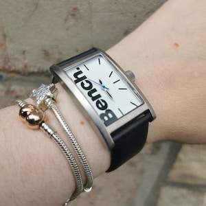 May include: A rectangular Bench watch with a white face, black hands, and a blue second hand. The watch has a silver-tone case and a black leather strap. The word "Bench" is printed on the watch face. A silver bracelet with charms is also visible.